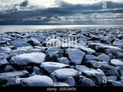 Kalte Winterlandschaft mit schneebedeckten Felsen neben einem großen Gewässer und stimmungsvollen Wolkenformationen. Stockfoto