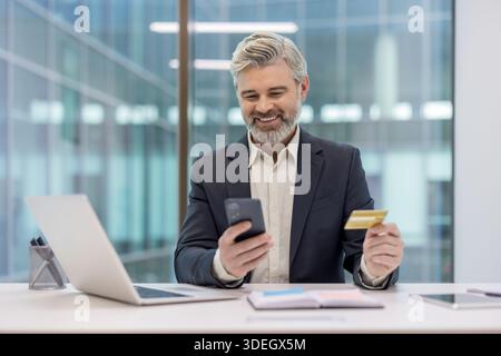 Lächelnder reifer Geschäftsmann am Schreibtisch mit Kreditkarte und Smartphone, während er eine Online-Zahlung oder einen E-Commerce-Kauf durchführt, selbstbewusst und entspannt beim Internet-Banking Stockfoto