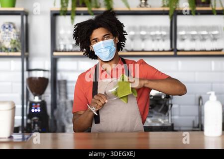 Arbeit Während Der Pandemie. Junger schwarzer Barkeeper mit medizinischer Maske, der an der Theke steht Stockfoto