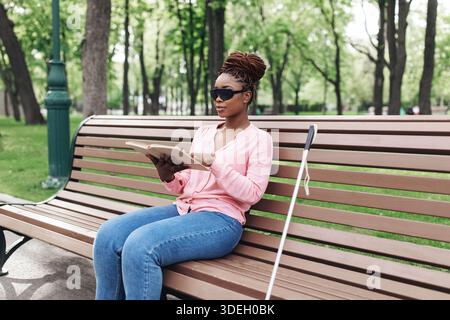 Junge schwarze Frau mit Sehbehinderung sitzt auf der Bank im Stadtpark, liest im Freien Braille-Buch, Kopierraum Stockfoto