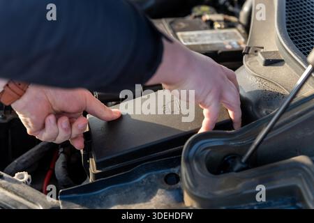 Die Abdeckung des Luftfilterkastens im Motorraum des Autos mit den Händen mit den Händen des Außenhändes, das staubige Kunststoffgehäuse und die Batterie sind im Hintergrund sichtbar. Konzept des Heimwerkerfahrzeugs Stockfoto