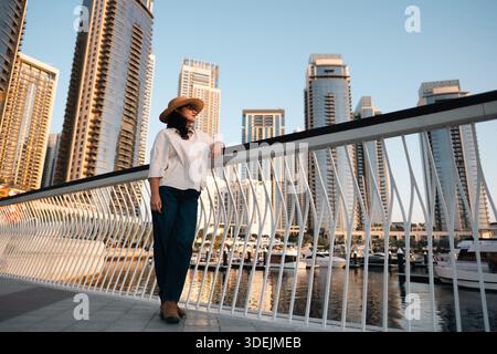 Selbstbewusste Frau, die an einem sonnigen Tag die Aussicht auf Dubai genießt, trägt einen Hut und ein elegantes Outfit, moderne Architektur im Hintergrund Stockfoto
