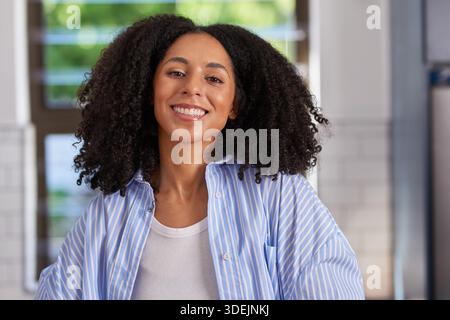Lächelnde Frau Mit Lockigem Haar In Gestreiftem Blauem Hemd In Heller Inneneinrichtung Stockfoto