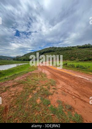 Eine gewundene rote Schotterstraße, die an einem ruhigen See und grünen Hügeln vorbeiführt, mit einem Motorrad in der Ferne im ländlichen Ruanda. Stockfoto
