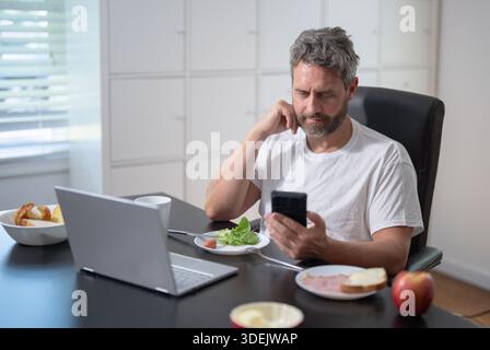Geschäftsmann isst morgens ein Essen, während er in einem gemütlichen Heimbüro seinen Laptop überprüft. Ein reifer Geschäftsmann, der seinen Arbeitstag plant. Ein Freelancer isst einen Stockfoto