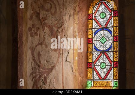 Das farbige Bleiglas einer romanischen Fensteröffnung wirft farbenfrohe Lichtreflexe auf die angrenzende Offenbarung mit floralen Wandgemälden im Inneren Stockfoto