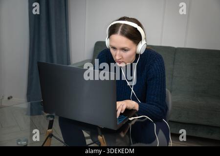 Junge Frau, die Kopfhörer mit Laptop trägt, während sie von zu Hause aus arbeitet und sich auf ihre Aufgaben in einer komfortablen und ungezwungenen Umgebung konzentriert Stockfoto