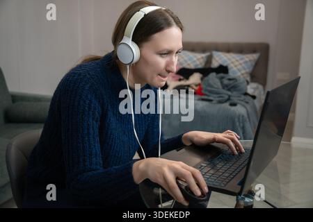 Junge Frau, die Kopfhörer trägt und Laptop benutzt, während sie von zu Hause aus arbeitet und sich trotz des unordentlichen Schlafzimmers auf ihre Aufgaben konzentriert Stockfoto