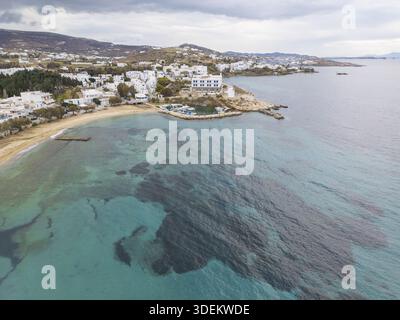 Die türkisfarbenen Wellen aus der Vogelperspektive umarmen sanft die Küste in der Nähe weiß getünchter Gebäude, im Kontrast zur zerklüfteten Küste Paros, Paros, Griechenland. Stockfoto