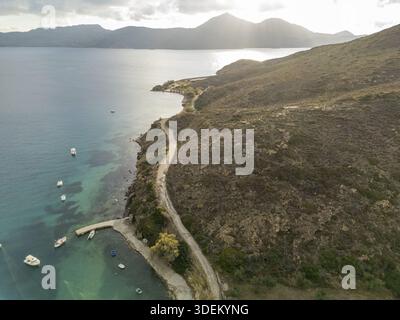 Aus der Vogelperspektive auf Boote, die sanft im türkisfarbenen Wasser in der Nähe der zerklüfteten Küste und der gewundenen Straße wippen, Adamantas, Milos, Griechenland. Stockfoto