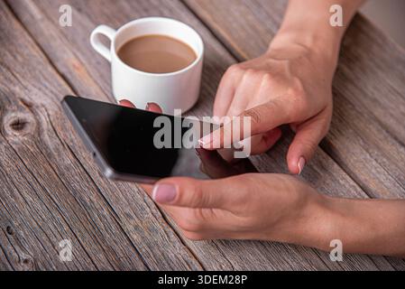 Die Hände einer Frau halten und interagieren mit einem schwarzen Smartphone auf einem rustikalen Holztisch Stockfoto