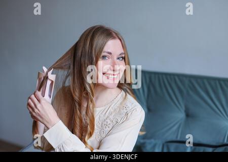Porträt einer fröhlichen Frau, die Haare im Wohnzimmer lockelt und die Schönheitspflege und das tägliche Wohlbefinden fördert Stockfoto