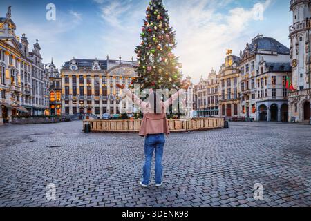 Eine glückliche Touristenfrau steht vor einem wunderschön geschmückten Weihnachtsbaum zur Weihnachtszeit am Grand Place in Brüssel, Belgien Stockfoto