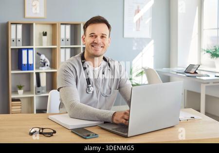 Porträt eines jungen glücklichen männlichen Arztes oder einer medizinischen Krankenschwester in Uniform, die am Schreibtisch in seinem Büro sitzt Stockfoto