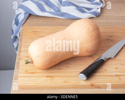 Ein ganzer Butternusskürbis liegt auf einem hölzernen Schneidebrett neben einem Messer, bereit zum Zerkleinern, Zubereiten von Mahlzeiten oder Kochen in der Küche Stockfoto
