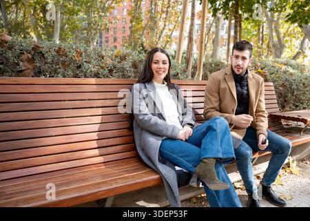 Zwei Freunde unterhalten sich auf einer Holzbank in einem pulsierenden Stadtpark, umgeben von farbenfrohen Herbstlaub und lebhafter Stadtlandschaft. Die goldene Sonne Stockfoto