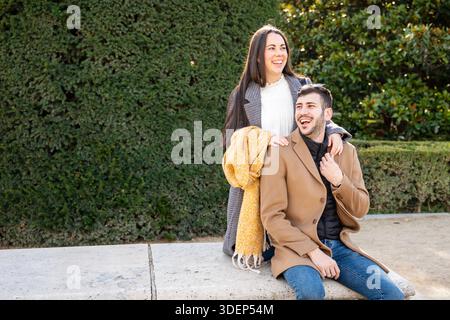 Zwei Freunde posieren glücklich in einem lebhaften Park, umgeben von Grün. Die Frau steht spielerisch hinter dem Mann, beide warm gekleidet für das kühle Wetter Stockfoto