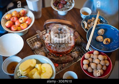 Blick von oben auf ein traditionelles chinesisches Teetreffen mit einem runden Holztisch, einer Holzkohle, einer Teekanne aus Glas und Schüsseln mit Obst, Nüssen, Süßkartoffeln, A Stockfoto