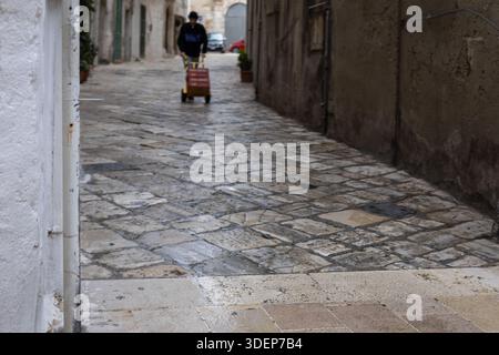 Ein Liefermann schiebt einen gelben Lastwagen mit Kisten durch eine enge, gepflasterte mediterrane Gasse, um das authentische tägliche Leben in einem alten europäischen Schlepptau festzuhalten Stockfoto
