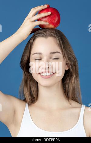 Eine junge Frau lächelt mit geschlossenen Augen und balanciert einen frischen Granatapfel auf dem Kopf Stockfoto