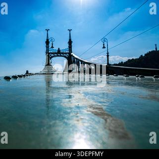 Flacher Blick auf eine historische europäische Brücke mit dekorativen Lampenpfosten, Reflexionen auf nasser Oberfläche und klarem blauem Himmel. Stockfoto