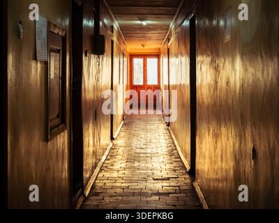 Langer, schmaler Korridor mit warmem Licht, das von Holzwänden und einem Backsteinboden reflektiert wird, führt zu einer hellen Ausgangstür, die Konzepte von PA vermittelt Stockfoto