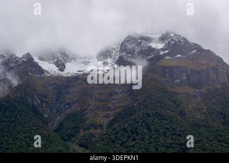 Eine dramatische Berglandschaft, umgeben von Wolken, mit schneebedeckten Gipfeln und üppigen grünen Wäldern am Fuß. Die Szene vermittelt ein Gefühl von Rätsel Stockfoto