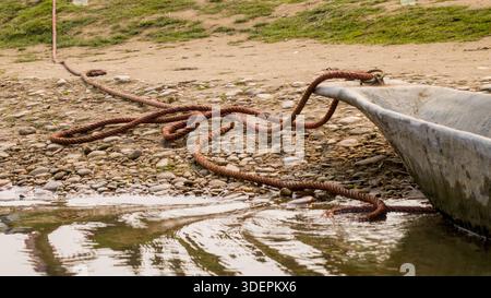 Ein traditionelles Holzboot liegt am felsigen Ufer, mit einem langen, losen Seil, das lässig auf dem Kieselboden liegt. Stockfoto