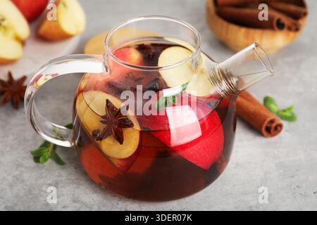 Aromatischer Früchtetee in Glas-Teekanne, Äpfel, Minze und Gewürze auf grauem Tisch, Nahaufnahme Stockfoto