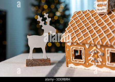 Weiße Hirschfigur aus Holz auf Rinde steht neben dem Lebkuchenhaus mit Glasur und Fensterlicht auf dem Tisch mit goldenem Bokeh des Weihnachtsbaums. Konzept von Stockfoto
