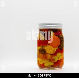 Ein Glasgefäß fasst eine Mischung aus bunten Gemüse und Früchten in einer Flüssigkeit. Der Becher wird auf eine weiße Oberfläche gestellt. Diese Szene eignet sich für die Lagerung und Zubereitung von Lebensmitteln. Stockfoto