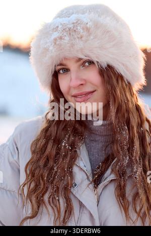 Dieses Bild fängt die heitere Schönheit einer jungen Frau in einer Winterlandschaft ein. Das Motiv, eine schöne Frau mit langen, lockigen braunen Haaren, steht im Mittelpunkt Stockfoto