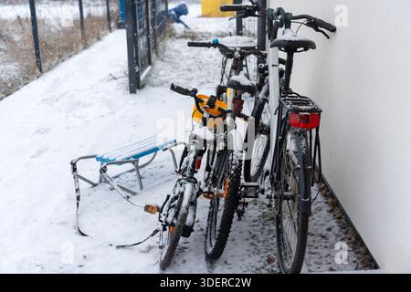 Schneebedeckte Fahrräder mit Kindersitz in der Nähe der weißen Wand mit Kinderschlitten auf verschneiten Boden im Innenhof während des Winters Schnee. Konzept von Stockfoto
