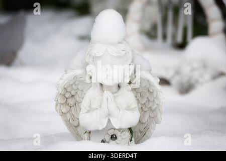 Nahaufnahme einer weißen Engelfigur aus dem Engel, die in einem verschneiten Garten betet. Ein Schneehaufen sitzt wie ein Hut auf dem Kopf. Friedliche Winterszene. Stockfoto