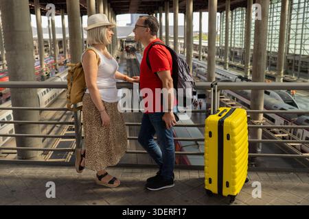 Glückliches reifes Touristenpaar, das in einem großen Bahnhof steht und ihre Reise mit Rucksäcken und einem gelben Koffer genießt. Stockfoto