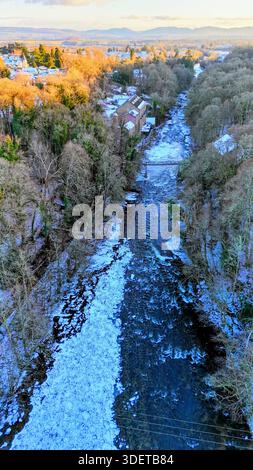 Aus der Vogelperspektive auf den frostbedeckten Fluss Ericht in der Nähe von Cargill’s Leap in Blairgowrie mit einer kleinen Brücke und einer alten Mühle am Fluss, umgeben von einer kalten Winterlandschaft Stockfoto