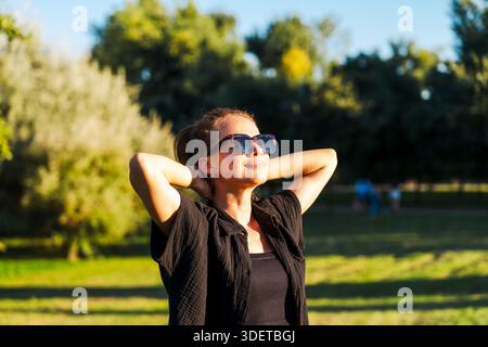Porträt einer Frau, die Hände hinter Kopf hält und die urbane Natur an sonnigen Tagen genießt. Stockfoto