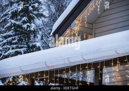 Nahaufnahme des schneebedeckten Daches und der Veranda mit warmen glühenden WeihnachtsLichtern am Winterhaus. Gemütliche Weihnachtsdekoration, Außenansicht der kalten Jahreszeit Stockfoto