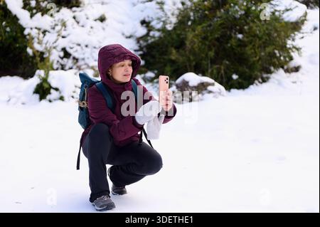 Eine Frau in kastanienbrauner Jacke und Rucksack hockt sich in einer verschneiten Landschaft und hält ein Smartphone, um ein Selfie zu machen. Handschuhe an, konzentriert auf den Bildschirm. Stockfoto