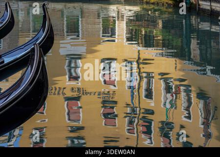 Die schwarzen Gondeln schweben auf einem ruhigen Kanal, wo sich die gelbe Gebäudefassade im plätschernden Wasser spiegelt, was ein verzerrtes Spiegelbild in Venedig, Italien, erzeugt. Stockfoto
