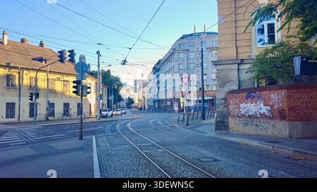 Eine geschäftige Stadtstraße zeigt Straßenbahnschienen, Verkehrsschilder, Autos und Fußgänger neben Graffitis-bedeckten Wänden und gemischter Architektur, die den Alltag einfangen Stockfoto