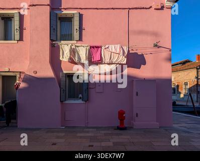 Burano, Venedig, Venetien, Italien. Die Wäsche hängt an der pinkfarbenen Wand. Sonnenlicht wirft scharfen Schatten auf die Fassade. Fenster unter der Linie geöffnet. Stockfoto