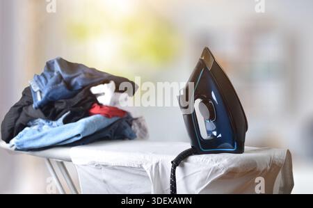 Konzept des Bügelens von Kleidung mit Bügeleisen auf einem Bügelbrett mit einem Stapel von Kleidung in einem Zimmer. Vorderansicht. Stockfoto