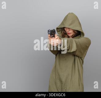 Selbstbewusste Frau im Kapuzenpullover, die Augen schließt, die Waffe in den Händen hält, bereit zum Schießen. Weibliche Sicherheitsbeamte, Gleichstellung der Geschlechter. Grauer Hintergrund. Stockfoto
