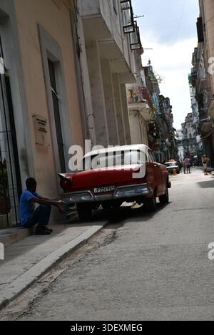 Mann sitzt neben einem geparkten Oldtimer in einer engen Straße in Old Havanna, Kuba Stockfoto