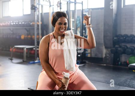 Afroamerikanerin sitzt auf einer Turnbank im pinkfarbenen Tank, hält die Wasserflasche und beugt den Arm Stockfoto