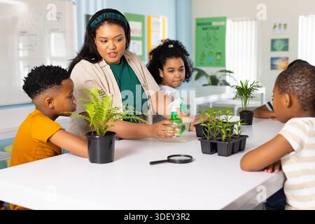 Afroamerikaner Lehrer mit drei Kindern, die Pflanzenpflege im Klassenzimmer zeigen und grüne Sprühflasche halten Stockfoto