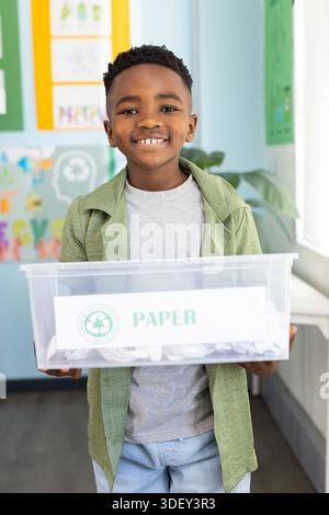 Afroamerikanisches Kind lächelt und hält im Klassenzimmer klares Plastikeimer mit PAPIER Stockfoto