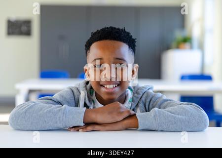 Afroamerikanischer Junge sitzt am weißen Klassentisch, lehnt sich auf die Arme und trägt einen grauen Hoodie Stockfoto