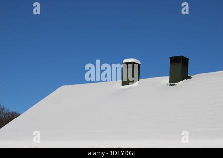 Schnee bedeckt das Dach eines Gebäudes mit zwei grünen Schornsteinen. Der hellblaue Himmel ist im Hintergrund sichtbar und zeigt einen klaren Wintertag. Stockfoto